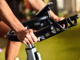 Close-up das mãos de uma pessoa utilizando uma bicicleta ergométrica em uma academia. A imagem destaca a pegada firme no guidão e o painel do equipamento ao fundo, representando um exercício cardiovascular de baixo impacto.