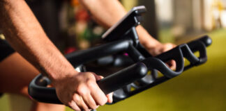 Close-up das mãos de uma pessoa utilizando uma bicicleta ergométrica em uma academia. A imagem destaca a pegada firme no guidão e o painel do equipamento ao fundo, representando um exercício cardiovascular de baixo impacto.