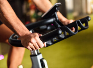 Close-up das mãos de uma pessoa utilizando uma bicicleta ergométrica em uma academia. A imagem destaca a pegada firme no guidão e o painel do equipamento ao fundo, representando um exercício cardiovascular de baixo impacto.