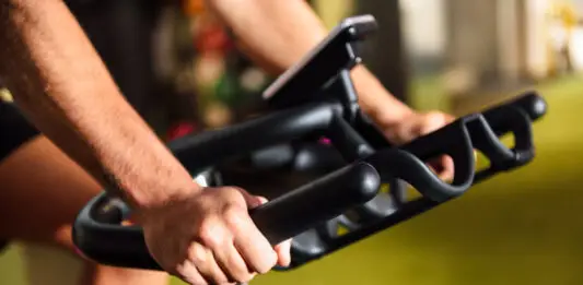 Close-up das mãos de uma pessoa utilizando uma bicicleta ergométrica em uma academia. A imagem destaca a pegada firme no guidão e o painel do equipamento ao fundo, representando um exercício cardiovascular de baixo impacto.