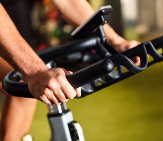 Close-up das mãos de uma pessoa utilizando uma bicicleta ergométrica em uma academia. A imagem destaca a pegada firme no guidão e o painel do equipamento ao fundo, representando um exercício cardiovascular de baixo impacto.