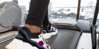 Close das pernas e dos pés de uma pessoa caminhando ou correndo em uma esteira, usando calça esportiva preta e tênis brancos com detalhes coloridos, em uma academia iluminada por grandes janelas.