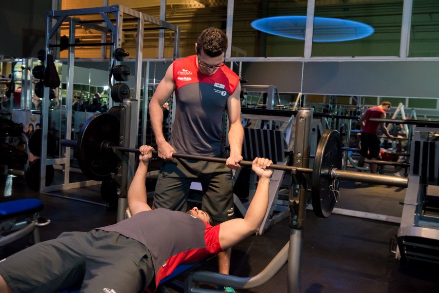 Professor auxiliando aluno no exercício supino reto com barra.