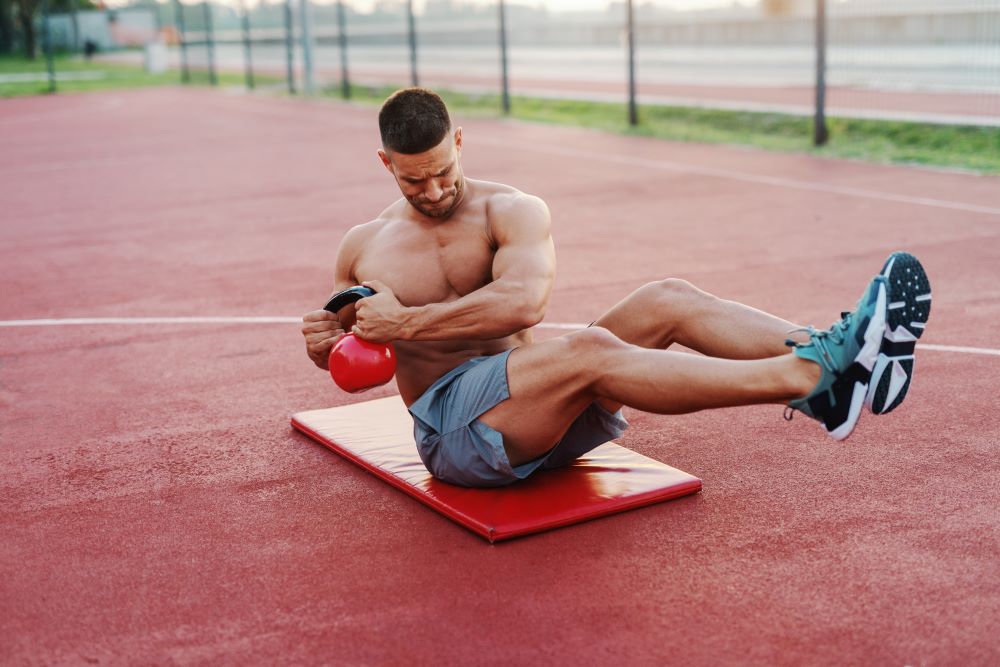 Homem sem camisa sentado em colchonete em quadra segurando kettlebell.