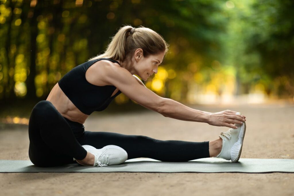 Mulher loira com roupa de ginástica preta sentada no chão de parque alongando as pernas.