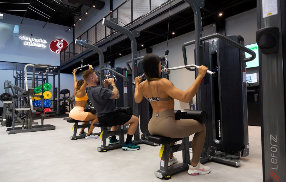 Estudantes de academia realizando exercício de puxada alta frontal em aparelho.