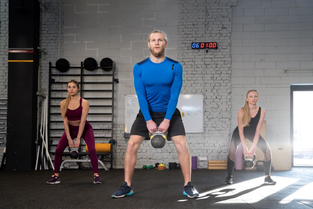 Alunos de academia com pernas afastadas e segurando kettlebell na frente para realizar exercício agachamento sumô.