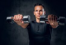 Homem segurando halteres na frente do corpo, na altura dos ombros, para realizar exercício de elevação frontal.