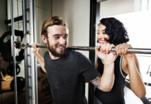 Homem realizando agachamento com barra em academia enquanto mulher o auxilia como instrutora.