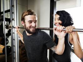 Homem realizando agachamento com barra em academia enquanto mulher o auxilia como instrutora.