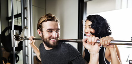 Exercícios de musculação: guia completo para iniciantes e intermediários Homem realizando agachamento com barra em academia enquanto mulher o auxilia como instrutora.