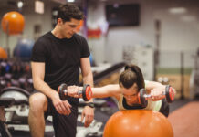 Mulher fazendo exercício com halteres sobre bola de estabilidade com acompanhamento de personal trainer na academia