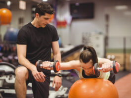 Mulher fazendo exercício com halteres sobre bola de estabilidade com acompanhamento de personal trainer na academia