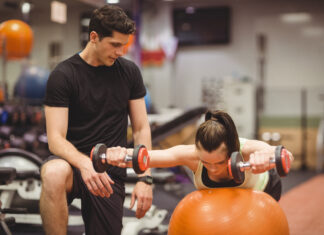 Mulher fazendo exercício com halteres sobre bola de estabilidade com acompanhamento de personal trainer na academia