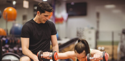 Mulher fazendo exercício com halteres sobre bola de estabilidade com acompanhamento de personal trainer na academia