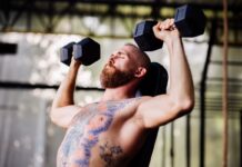 Homem tatuado fazendo desenvolvimento Arnold na academia.