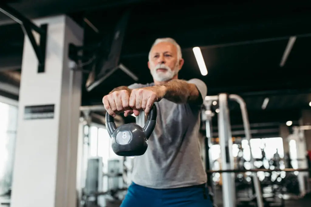 Homem idoso treina com kettlebell em academia moderna, realizando exercício de musculação com os braços estendidos à frente do corpo.