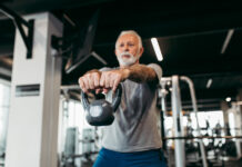 Homem idoso treina com kettlebell em academia moderna, realizando exercício de musculação com os braços estendidos à frente do corpo.