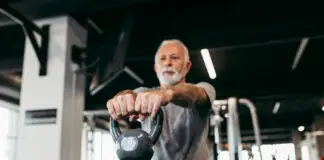 Homem idoso treina com kettlebell em academia moderna, realizando exercício de musculação com os braços estendidos à frente do corpo.