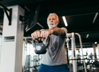 Homem idoso treina com kettlebell em academia moderna, realizando exercício de musculação com os braços estendidos à frente do corpo.