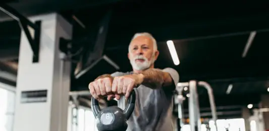 Homem idoso treina com kettlebell em academia moderna, realizando exercício de musculação com os braços estendidos à frente do corpo.