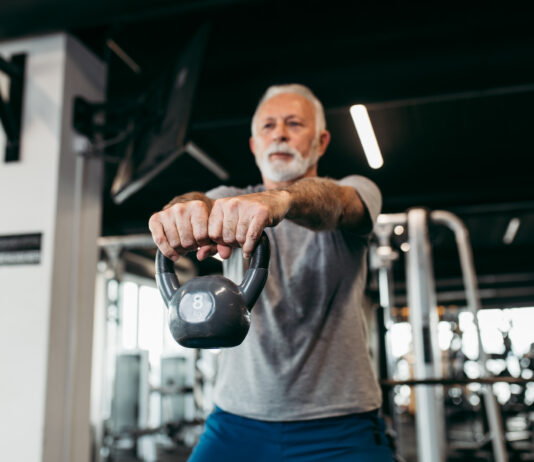 Homem idoso treina com kettlebell em academia moderna, realizando exercício de musculação com os braços estendidos à frente do corpo.