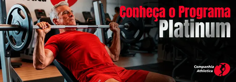 Homem idoso de cabelos grisalhos e camiseta vermelha praticando musculação no supino com barra em academia da Cia Athletica, dentro do Programa Platinum

