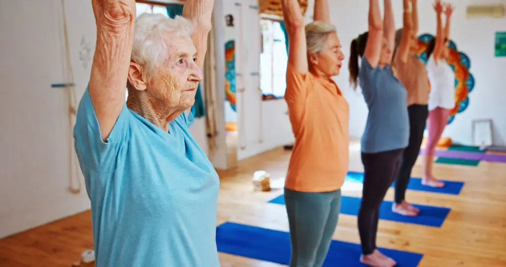 Grupo de mulheres idosas praticando Pilates em estúdio iluminado, com braços erguidos acima da cabeça durante exercício de alongamento e equilíbrio. Em primeiro plano, uma senhora de cabelos brancos usa camiseta azul-clara e demonstra concentração enquanto acompanha a aula. Ao fundo, outras alunas realizam o mesmo movimento sobre tapetes azuis em ambiente acolhedor e moderno.