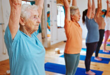 Grupo de mulheres idosas praticando Pilates em estúdio iluminado, com braços erguidos acima da cabeça durante exercício de alongamento e equilíbrio. Em primeiro plano, uma senhora de cabelos brancos usa camiseta azul-clara e demonstra concentração enquanto acompanha a aula. Ao fundo, outras alunas realizam o mesmo movimento sobre tapetes azuis em ambiente acolhedor e moderno.