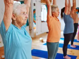 Grupo de mulheres idosas praticando Pilates em estúdio iluminado, com braços erguidos acima da cabeça durante exercício de alongamento e equilíbrio. Em primeiro plano, uma senhora de cabelos brancos usa camiseta azul-clara e demonstra concentração enquanto acompanha a aula. Ao fundo, outras alunas realizam o mesmo movimento sobre tapetes azuis em ambiente acolhedor e moderno.