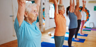 Grupo de mulheres idosas praticando Pilates em estúdio iluminado, com braços erguidos acima da cabeça durante exercício de alongamento e equilíbrio. Em primeiro plano, uma senhora de cabelos brancos usa camiseta azul-clara e demonstra concentração enquanto acompanha a aula. Ao fundo, outras alunas realizam o mesmo movimento sobre tapetes azuis em ambiente acolhedor e moderno.