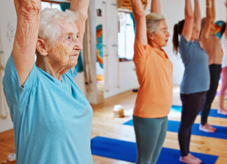 Grupo de mulheres idosas praticando Pilates em estúdio iluminado, com braços erguidos acima da cabeça durante exercício de alongamento e equilíbrio. Em primeiro plano, uma senhora de cabelos brancos usa camiseta azul-clara e demonstra concentração enquanto acompanha a aula. Ao fundo, outras alunas realizam o mesmo movimento sobre tapetes azuis em ambiente acolhedor e moderno.