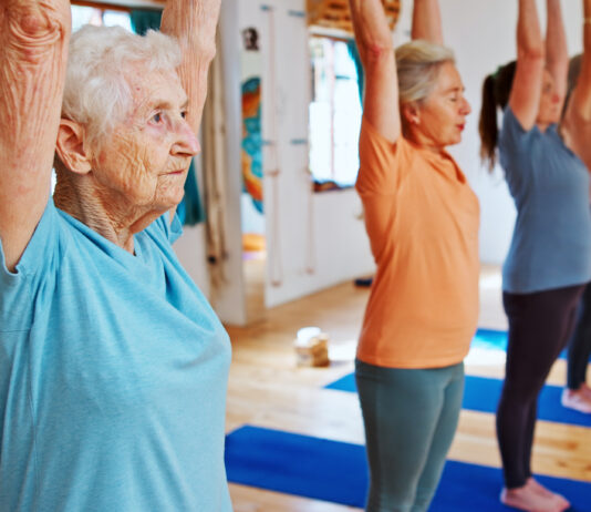 Grupo de mulheres idosas praticando Pilates em estúdio iluminado, com braços erguidos acima da cabeça durante exercício de alongamento e equilíbrio. Em primeiro plano, uma senhora de cabelos brancos usa camiseta azul-clara e demonstra concentração enquanto acompanha a aula. Ao fundo, outras alunas realizam o mesmo movimento sobre tapetes azuis em ambiente acolhedor e moderno.