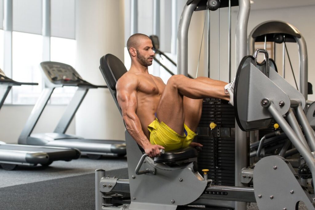 Um homem musculoso executa o Leg Press Horizontal em uma máquina de musculação para pernas, em uma academia bem iluminada. Ele está com os joelhos flexionados, usando as alças laterais para estabilizar o tronco.