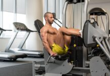Um homem musculoso executa o Leg Press Horizontal em uma máquina de musculação para pernas, em uma academia bem iluminada. Ele está com os joelhos flexionados, usando as alças laterais para estabilizar o tronco.