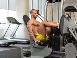 Leg press horizontal: guia completo para ganhar força e definição muscular Um homem musculoso executa o Leg Press Horizontal em uma máquina de musculação para pernas, em uma academia bem iluminada. Ele está com os joelhos flexionados, usando as alças laterais para estabilizar o tronco.