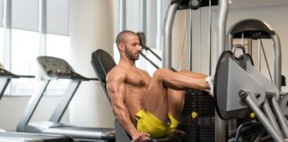 Um homem musculoso executa o Leg Press Horizontal em uma máquina de musculação para pernas, em uma academia bem iluminada. Ele está com os joelhos flexionados, usando as alças laterais para estabilizar o tronco.