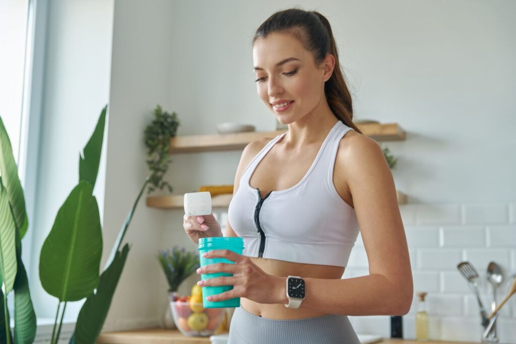 A imagem mostra uma mulher sorridente com roupas de ginástica, em uma cozinha bem iluminada, preparando um suplemento em um shaker azul. A cena, com frutas e plantas ao fundo, sugere um estilo de vida focado em saúde e bem-estar.