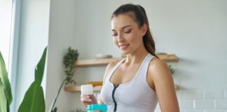 A imagem mostra uma mulher sorridente com roupas de ginástica, em uma cozinha bem iluminada, preparando um suplemento em um shaker azul. A cena, com frutas e plantas ao fundo, sugere um estilo de vida focado em saúde e bem-estar.