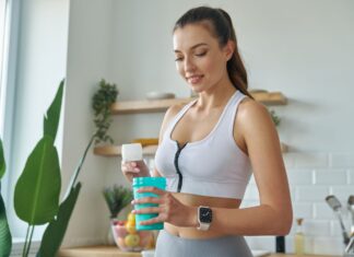 Como tomar creatina: dosagem, horário e benefícios reais A imagem mostra uma mulher sorridente com roupas de ginástica, em uma cozinha bem iluminada, preparando um suplemento em um shaker azul. A cena, com frutas e plantas ao fundo, sugere um estilo de vida focado em saúde e bem-estar.