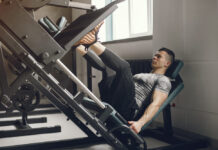 Erros no leg press 90 que podem travar seus resultados (e como corrigir) A imagem mostra um praticante de academia utilizando um aparelho de leg press 90°. A foto é capturada de cima, revelando principalmente as pernas do usuário posicionadas sobre a plataforma metálica inclinada. Ele veste tênis esportivos azuis com detalhes em verde e branco, além de shorts claros. A plataforma do equipamento possui textura antiderrapante e está conectada a barras laterais com anilhas carregadas, indicando que há peso sendo movimentado. As mãos do praticante seguram as alças laterais do assento para estabilização durante o exercício. O ambiente ao redor apresenta piso em tom arroxeado típico de áreas internas de academias. A cena transmite a execução do exercício de força focado nos membros inferiores, especialmente quadríceps, glúteos e posteriores de coxa