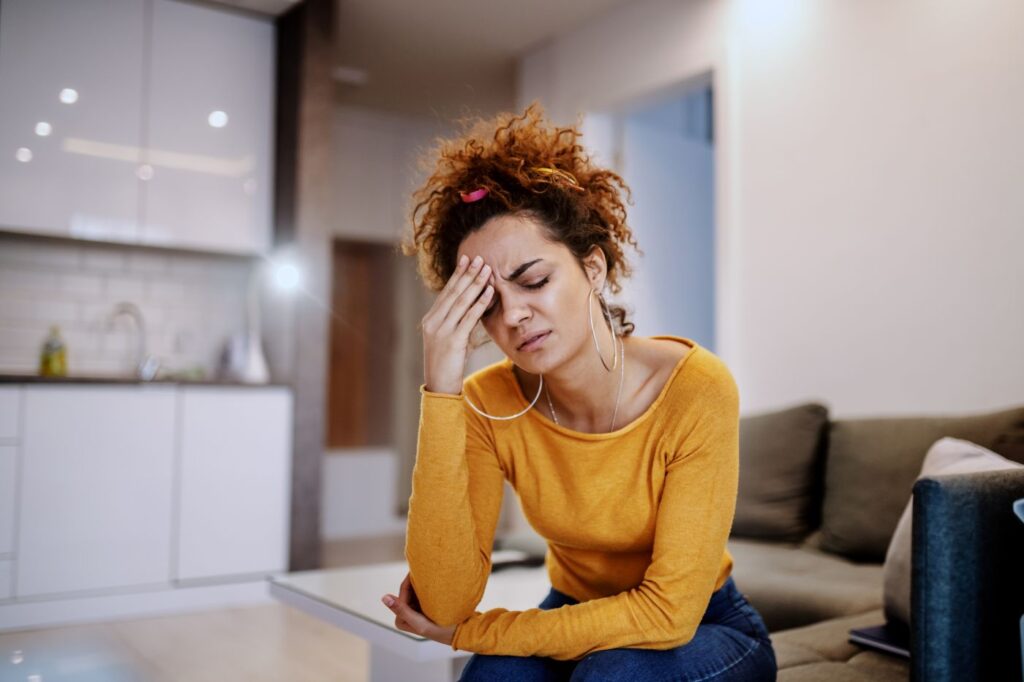 Mulher jovem com expressão de preocupação sentada no sofá, representando estresse e necessidade de aprender como diminuir a ansiedade com exercícios e respiração.