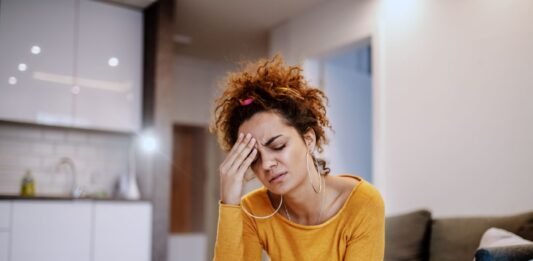 Como diminuir a ansiedade: 5 exercícios para acalmar a mente Mulher jovem com expressão de preocupação sentada no sofá, representando estresse e necessidade de aprender como diminuir a ansiedade com exercícios e respiração.