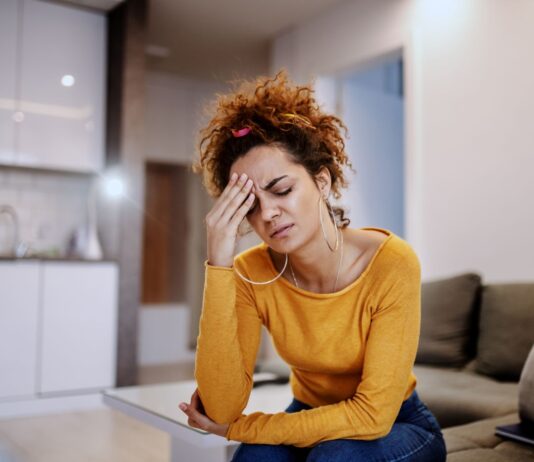 Como aliviar o estresse: 7 estratégias eficazes com exercícios Mulher jovem com expressão de preocupação sentada no sofá, representando estresse e necessidade de aprender como diminuir a ansiedade com exercícios e respiração.