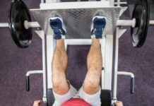 Pessoa realizando exercício no leg press 90 em academia, com pés na plataforma e pesos laterais, trabalhando músculos das pernas.