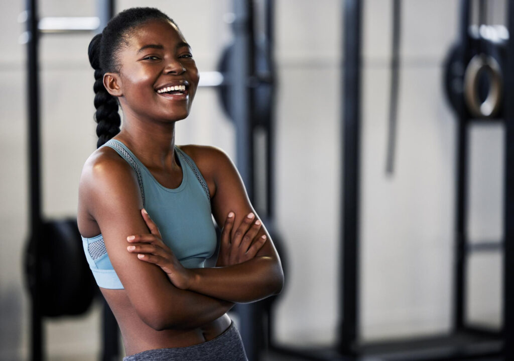 Mulher jovem e negra em ambiente de academia, sorrindo com os braços cruzados sinalizando a importância de exercícios físicos no combate aos sintomas da crise de ansiedade.