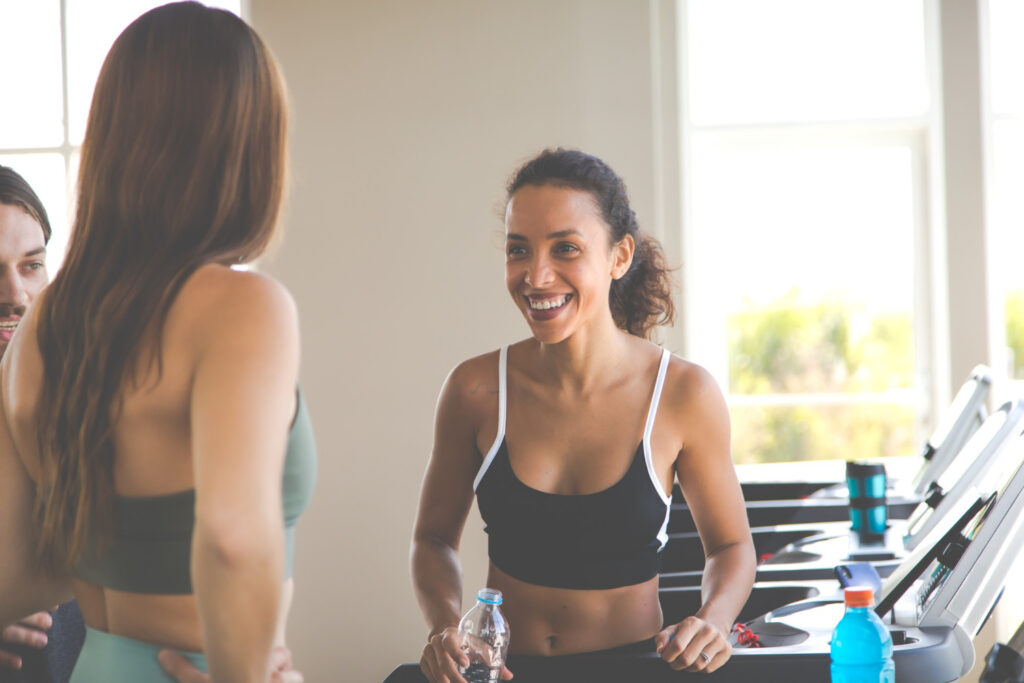 Mulheres interagindo na academia com garrafas de água e esteiras ao fundo - vida social ativa e saudável

