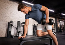 Homem executando remada unilateral com halter em banco na academia - remada com halteres