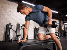 Remada com halteres: o movimento que trabalha muito mais do que as costas Homem executando remada unilateral com halter em banco na academia - remada com halteres