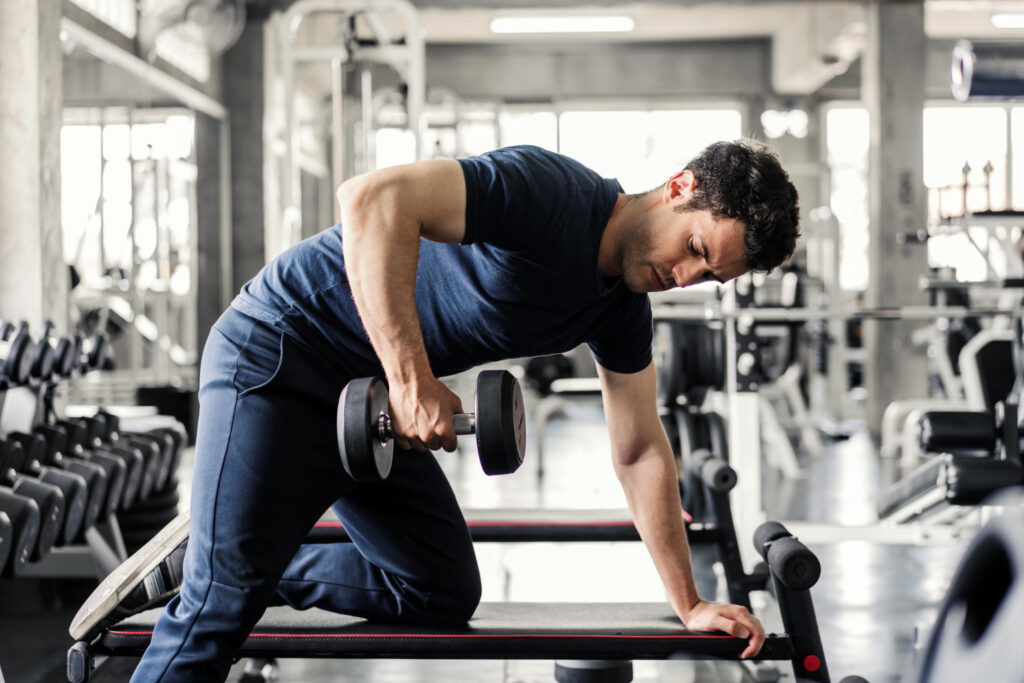 Homem executando remada unilateral com halter apoiado em banco dentro da academia